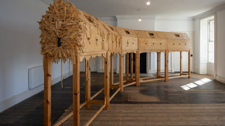 A wooden tunnel shaped sculpture on legs, curving around two walls of an upstairs room, with light streaming through the window on to the floor.